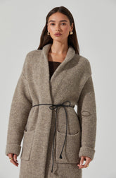 A woman is wearing a long, oversized, grey wool coat with a wide shawl collar. The coat has large front patch pockets and is cinched at the waist with a thin black drawstring. The sleeves are long and slightly rolled up at the cuffs. She is also wearing a black top underneath the coat. She has gold hoop earrings and rings on her fingers.